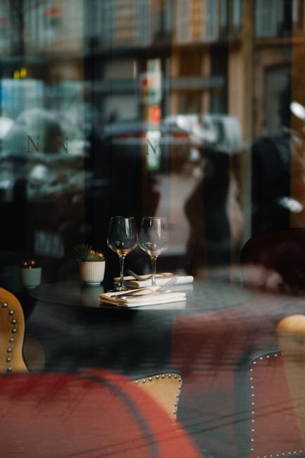 Elegantly set table in a city cafe, viewed through a window, featuring wine glasses and reflections.