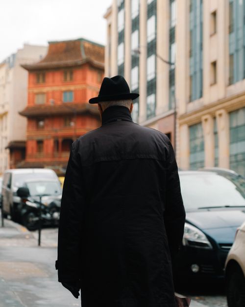 Free Man in black coat and hat walks past historic building in city street. Stock Photo