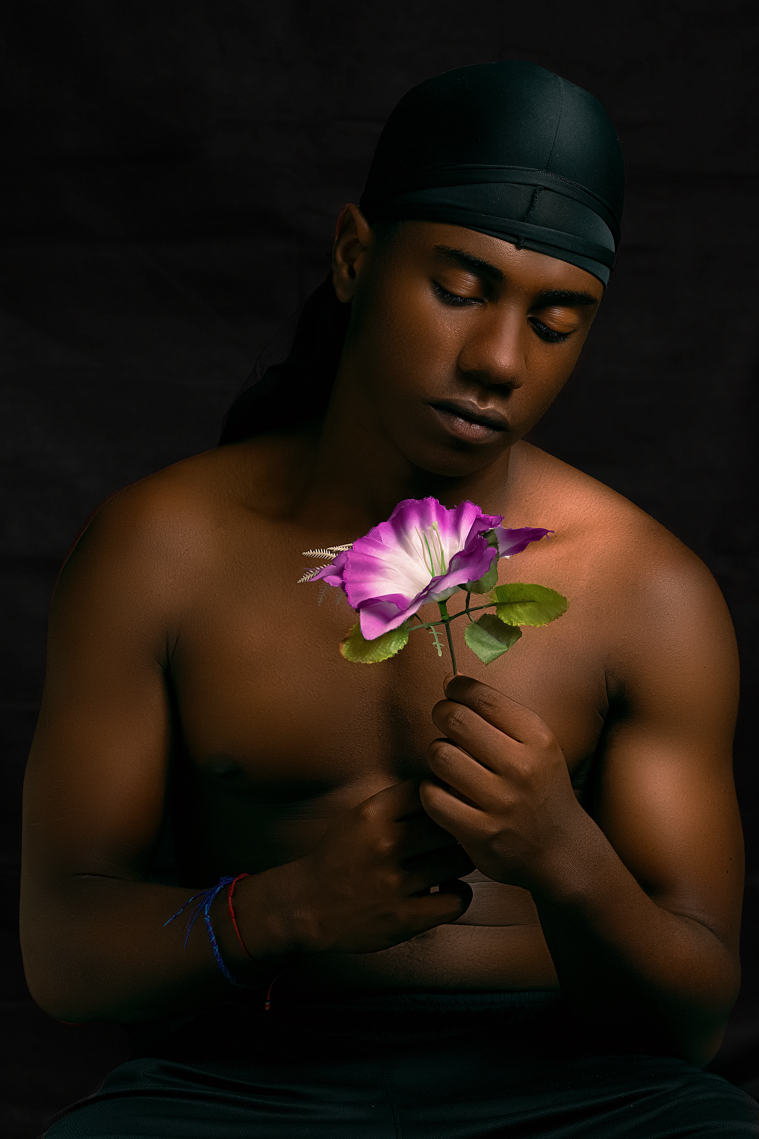 A topless man poses with a vibrant flower against a black backdrop, focusing on serenity and reflection.