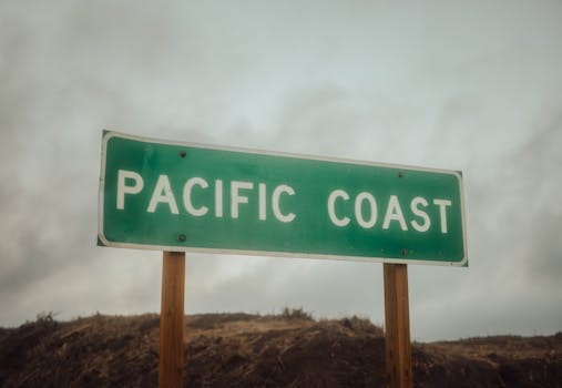 Green 'Pacific Coast' road sign against a dramatic overcast sky, evoking a sense of travel and adventure.