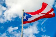 A vibrant Puerto Rican flag waving on a flagpole against a bright blue sky with clouds.