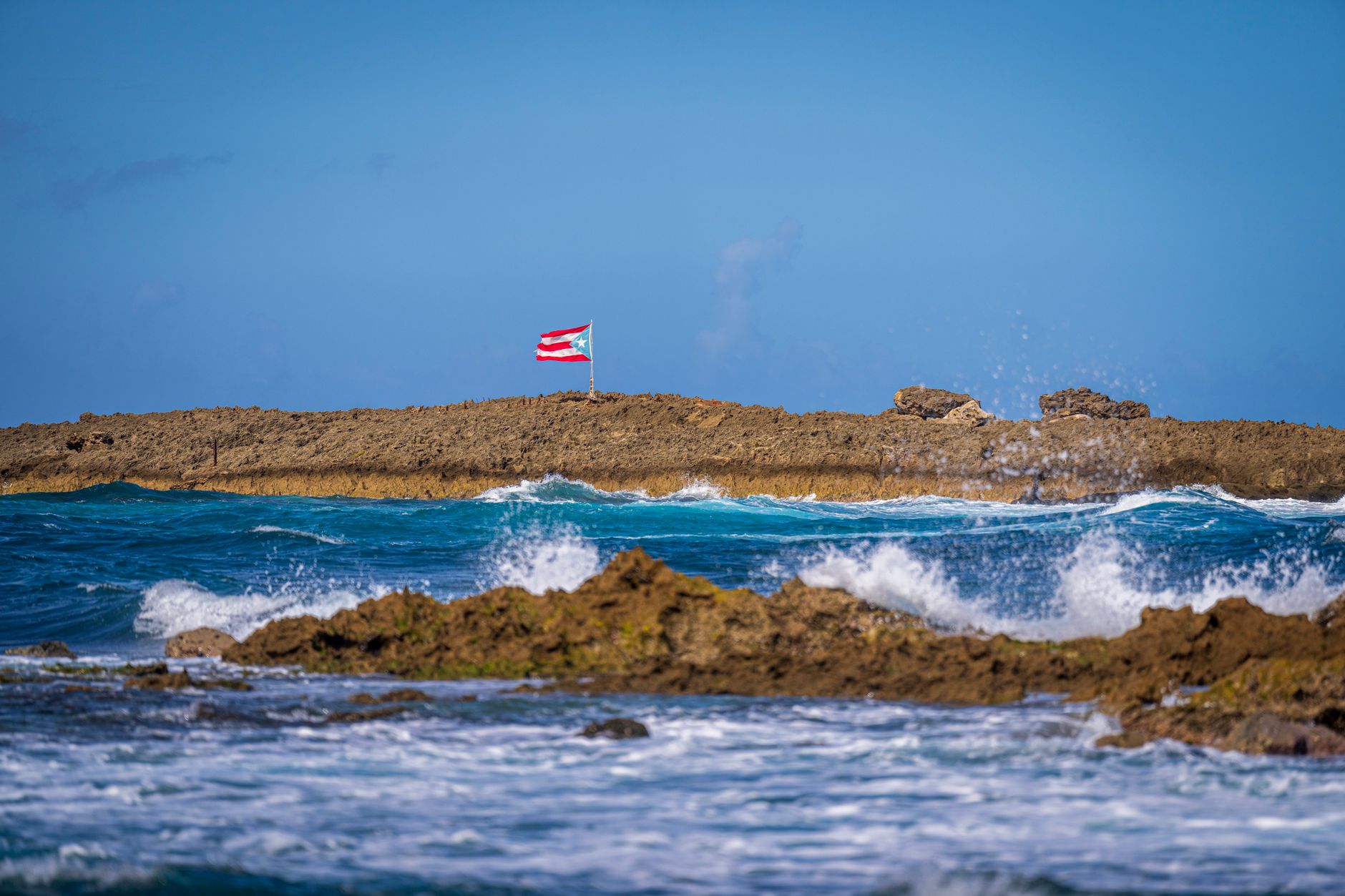 Photograph showcases the Puerto Rican flag waving on a rocky coastline against a clear blue sky.