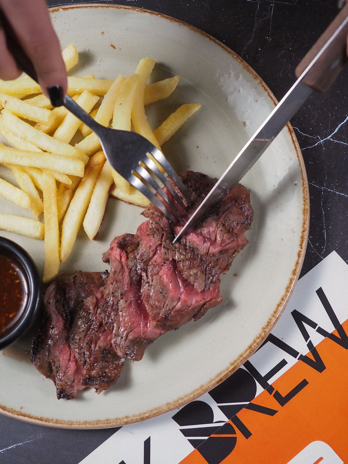 Overhead close-up of a plate with sliced medium-rare steak, golden shoestring fries and a small ramekin of dark sauce, fork and knife cutting into the meat