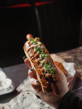 Close-up of a hand holding a gourmet hot dog with toppings in a dimly lit setting.