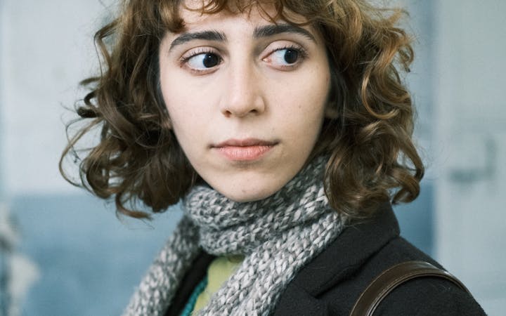 Portrait of a young woman with curly hair wearing warm clothing and a scarf indoors.
