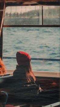 A woman with a red hat looks at the ocean through a ferry window, enjoying a peaceful journey.