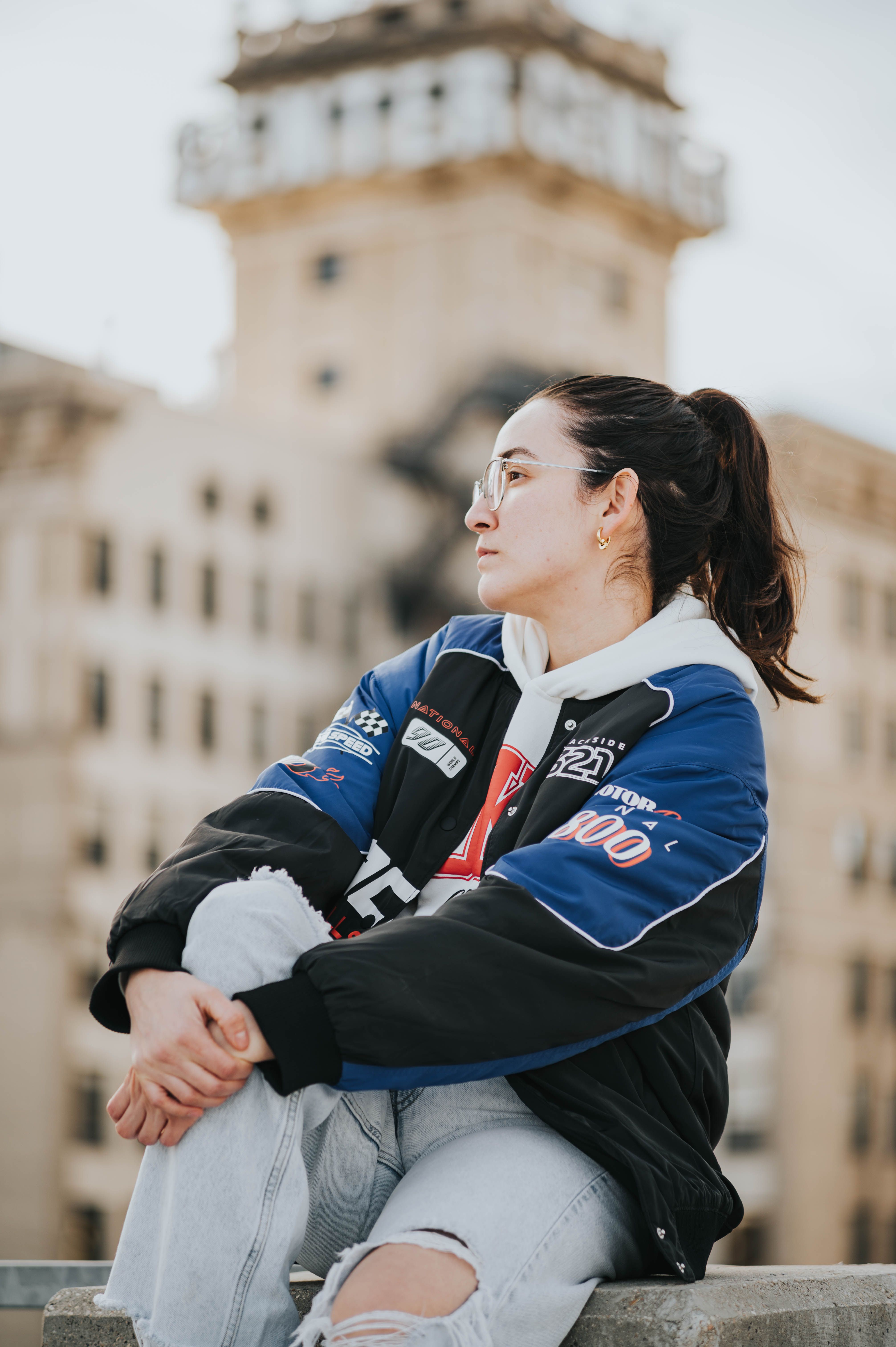 Stylish woman in trendy jacket sitting against an urban building backdrop, exuding youthful fashion vibes.