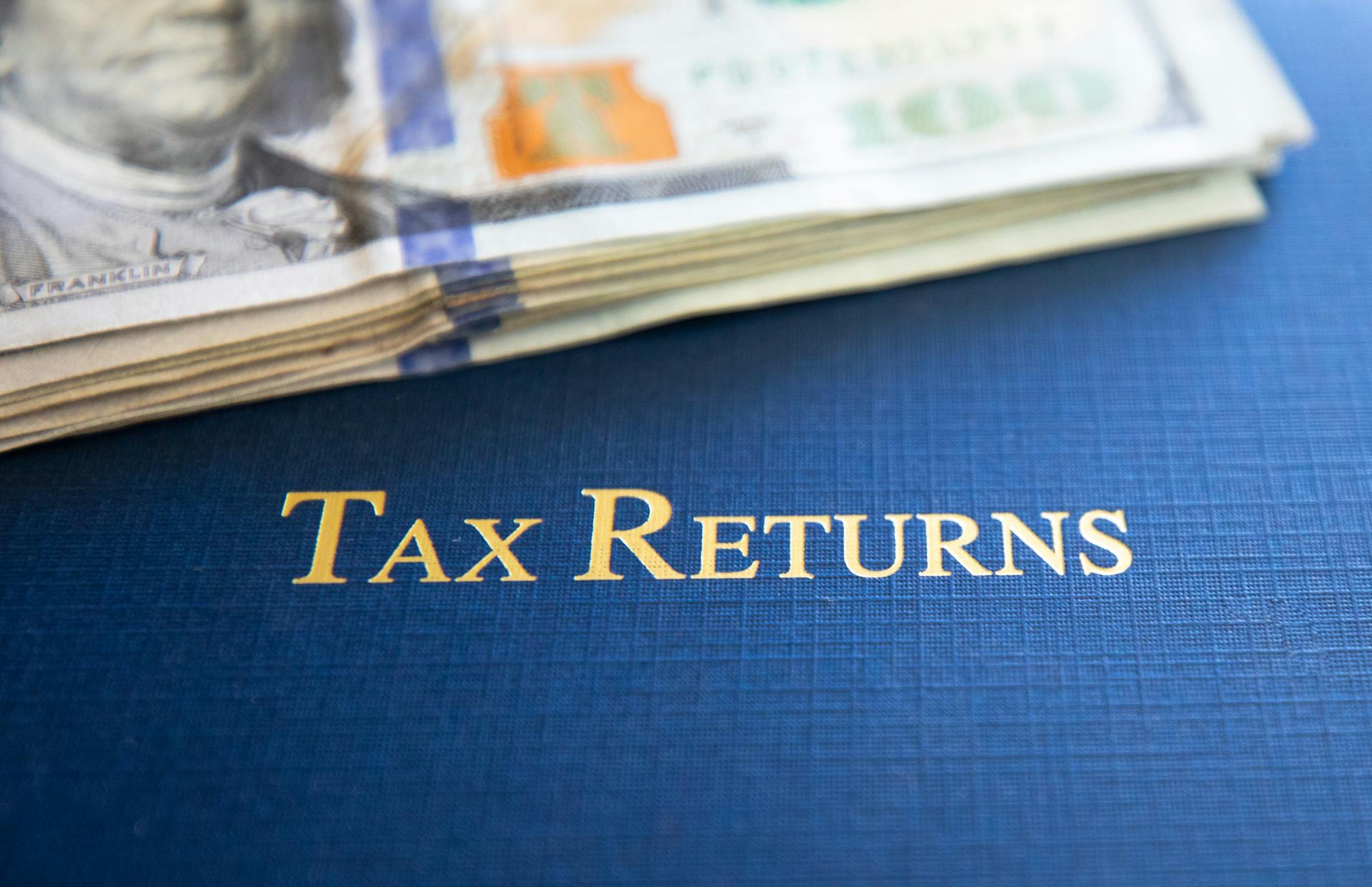 Close-up of tax returns folder beside a stack of American dollars, representing finance and accounting.