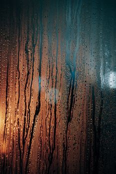 Raindrop-covered window with colorful reflections in a dark, moody setting.