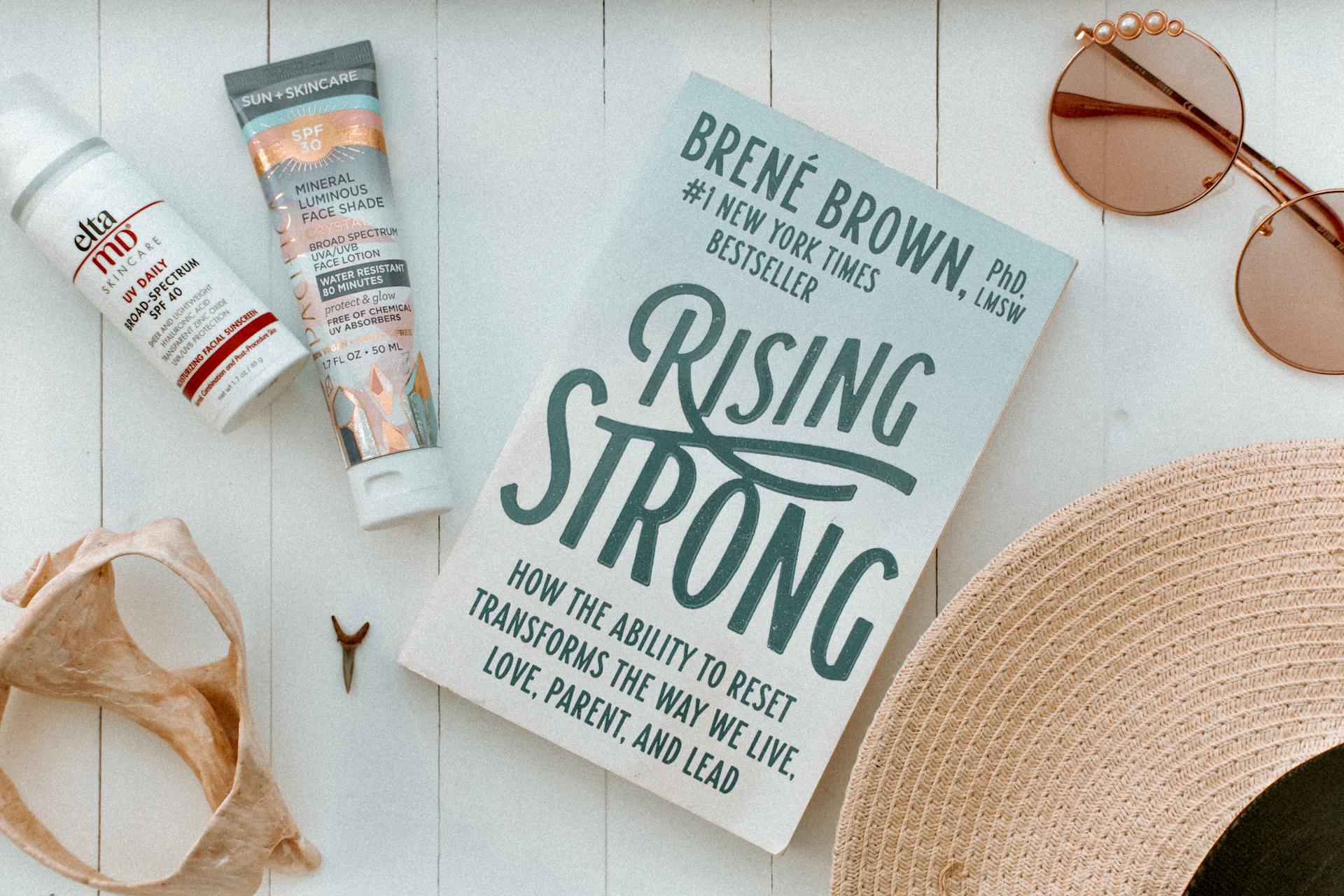 Flat lay of summer essentials including a book, SPF skincare, sunglasses, and a straw hat.