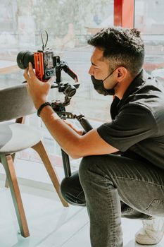 A photographer wearing a face mask uses a DSLR camera indoors with natural light.