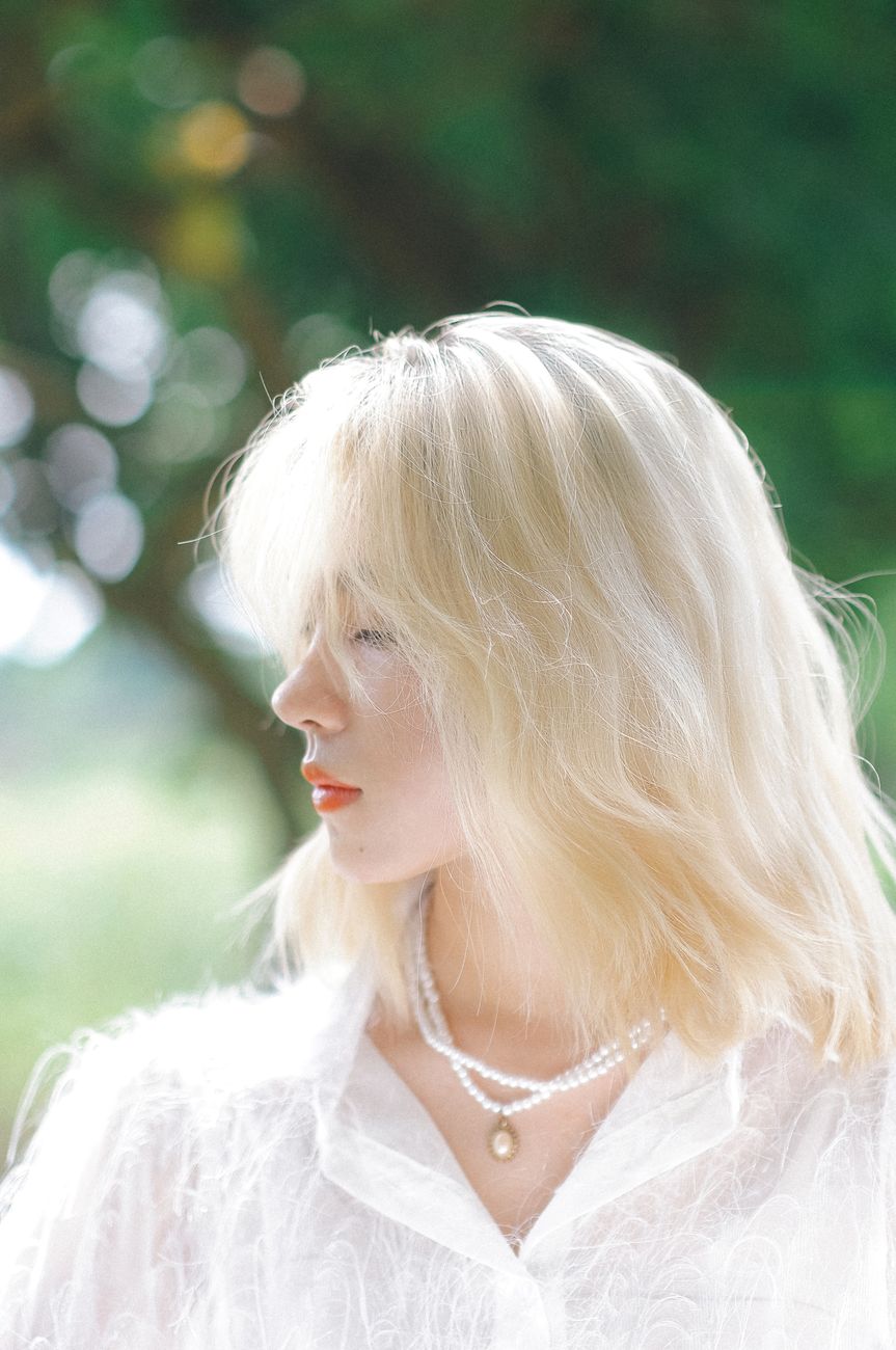 Profile portrait of a blonde woman wearing pearls against a green outdoor background.