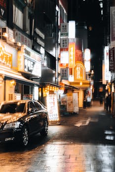 Illuminated street in bustling Asian city with neon signs and parked cars at night.