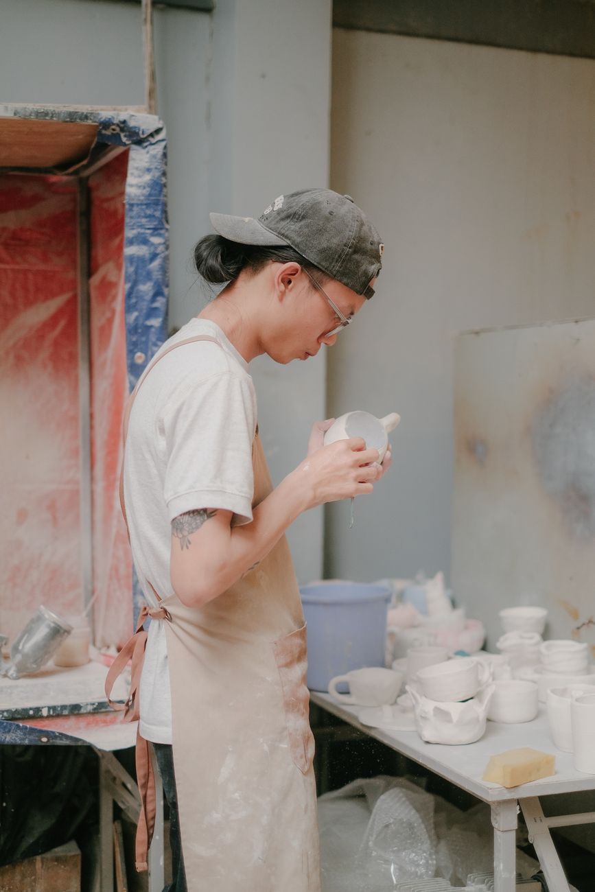 A young adult potter examines a ceramic cup in an indoor pottery studio setting.