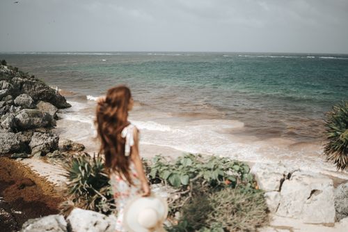 Free A woman with flowing brown hair stands at a rocky coast, gazing at the sea under an overcast sky. Stock Photo
