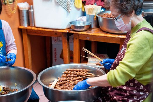 Elderly woman in apron preparing meat skewers in an indoor setting with large bowls.