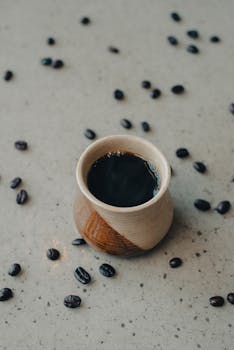 A rustic ceramic mug filled with black coffee surrounded by scattered coffee beans on a speckled surface.