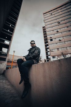Stylish man in sunglasses sitting on a wall in a modern urban setting.