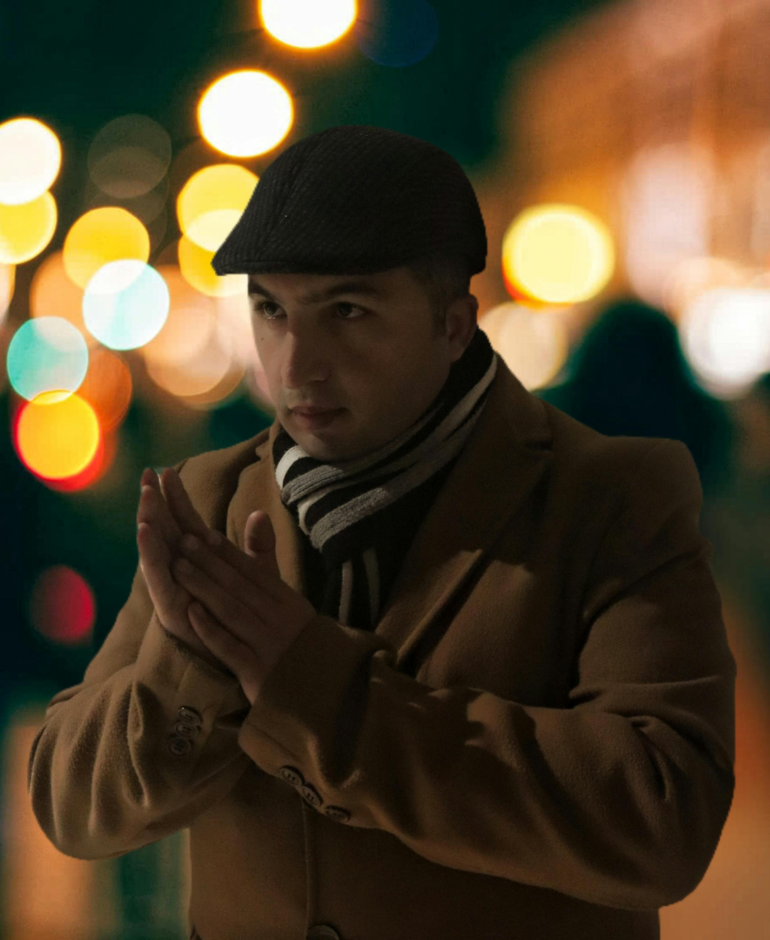 A stylish man in a coat and flat cap poses with a bokeh-filled city backdrop at night.