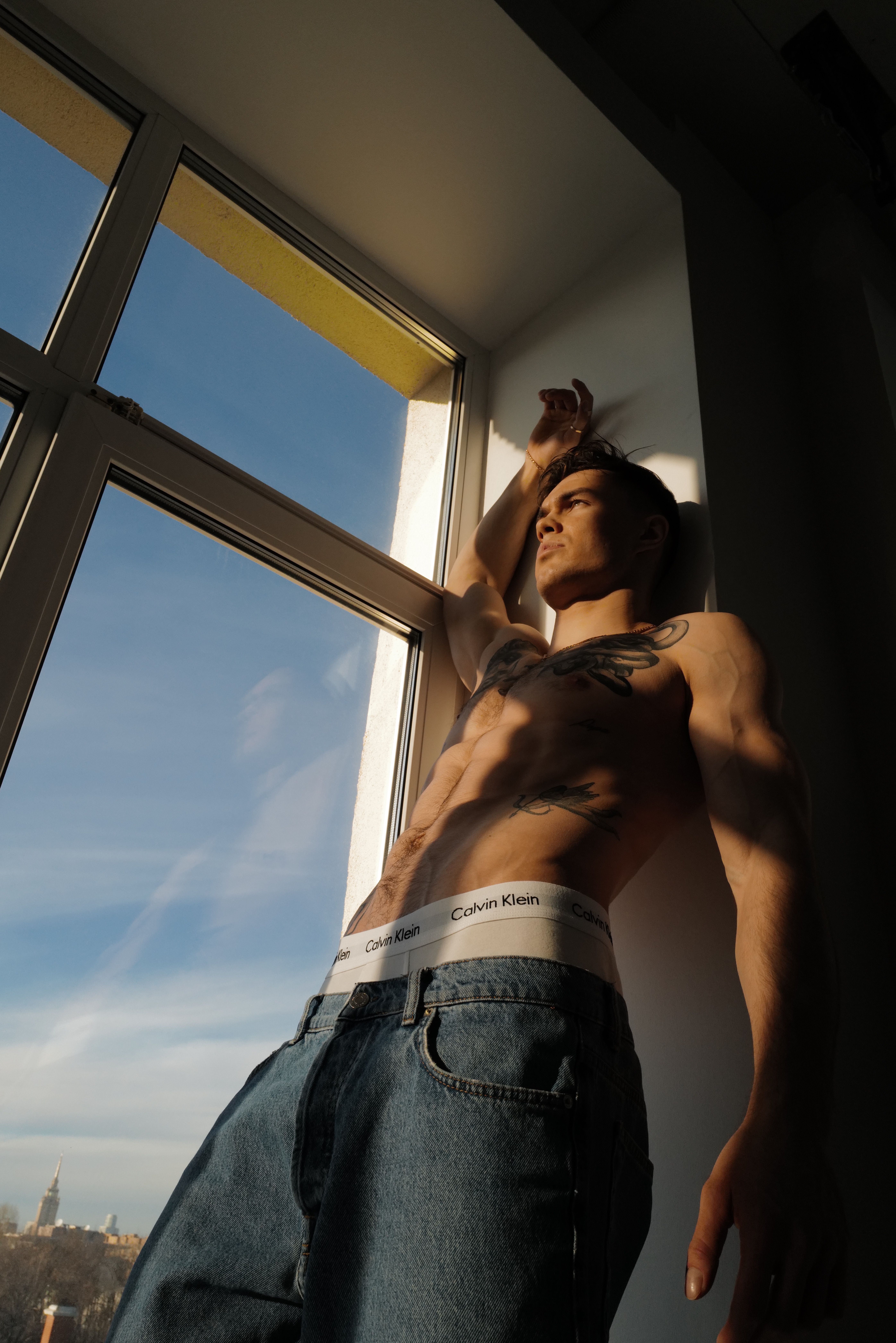 Tattooed man with muscular build posing topless by a window, showcasing a dynamic stance.
