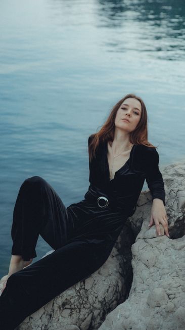 A woman in a black outfit poses gracefully on rocks by the ocean.