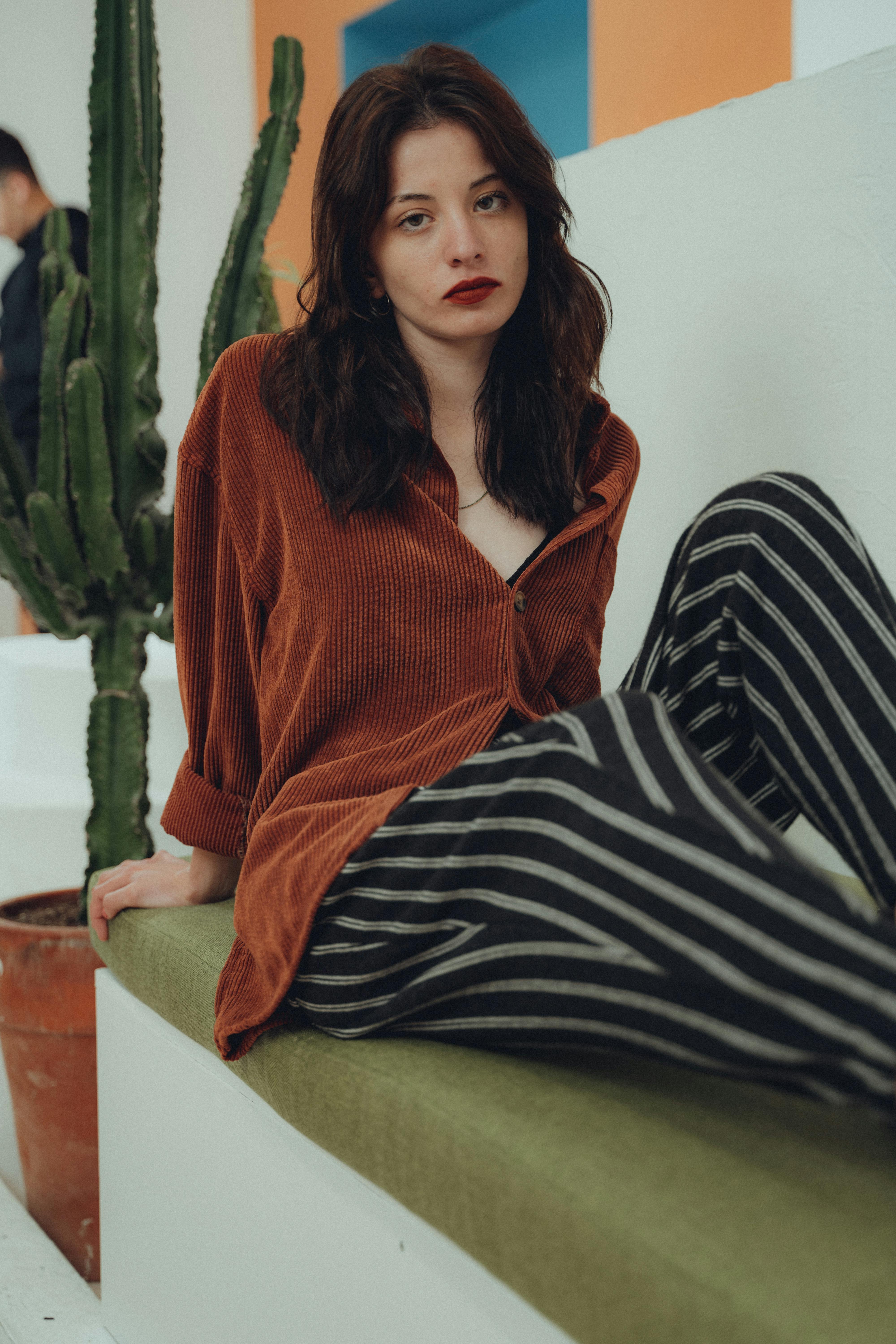 Stylish woman with long brown hair posing indoors beside a cactus plant.