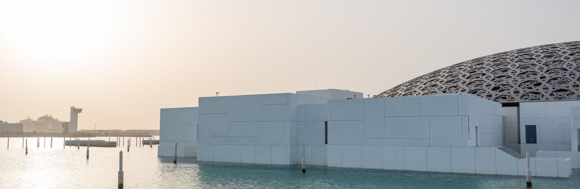 Futuristic white building with dome reflected in water at sunset, serene ambiance.
