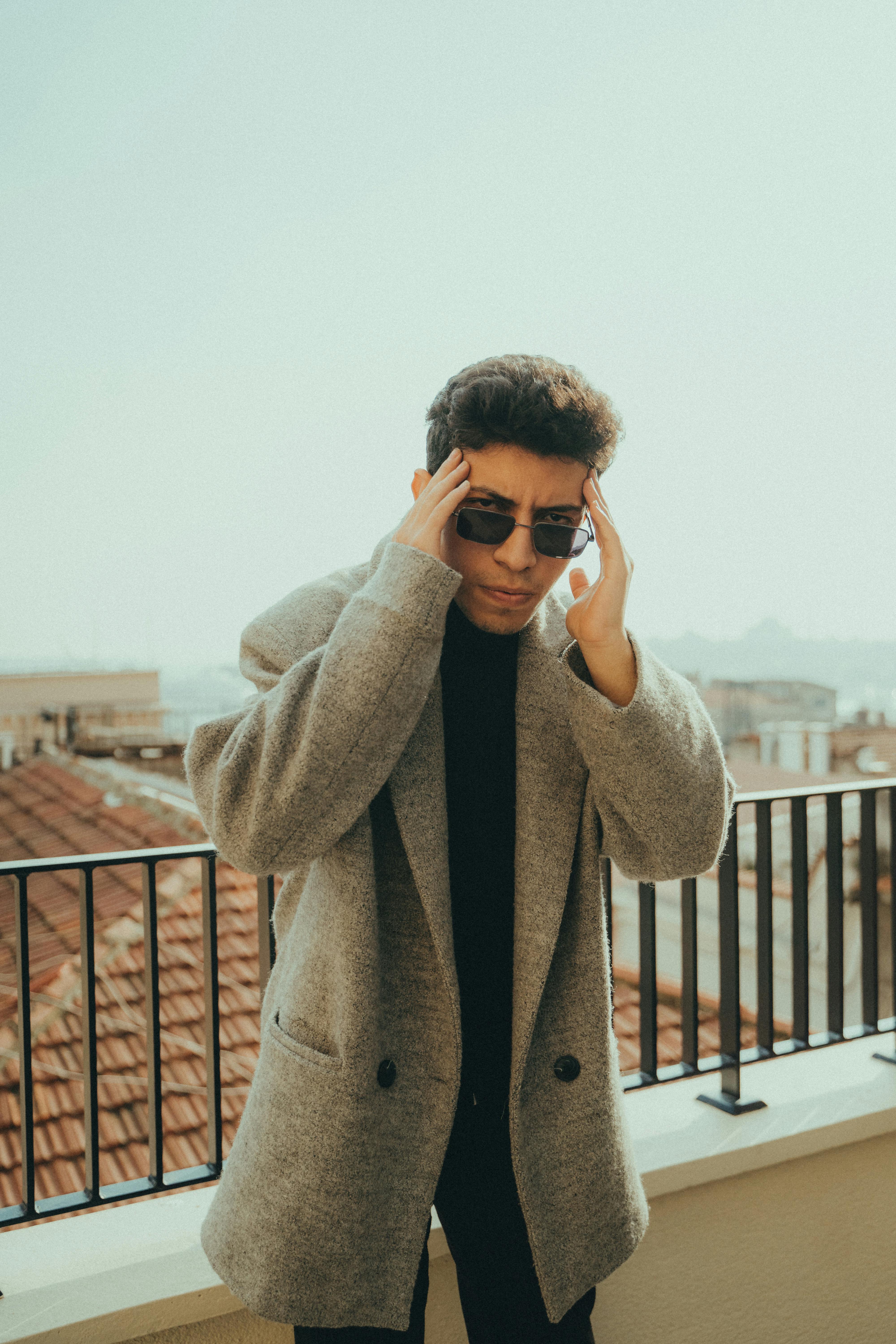 Trendy young man in sunglasses and coat posing on a rooftop with city view, embodying modern fashion.