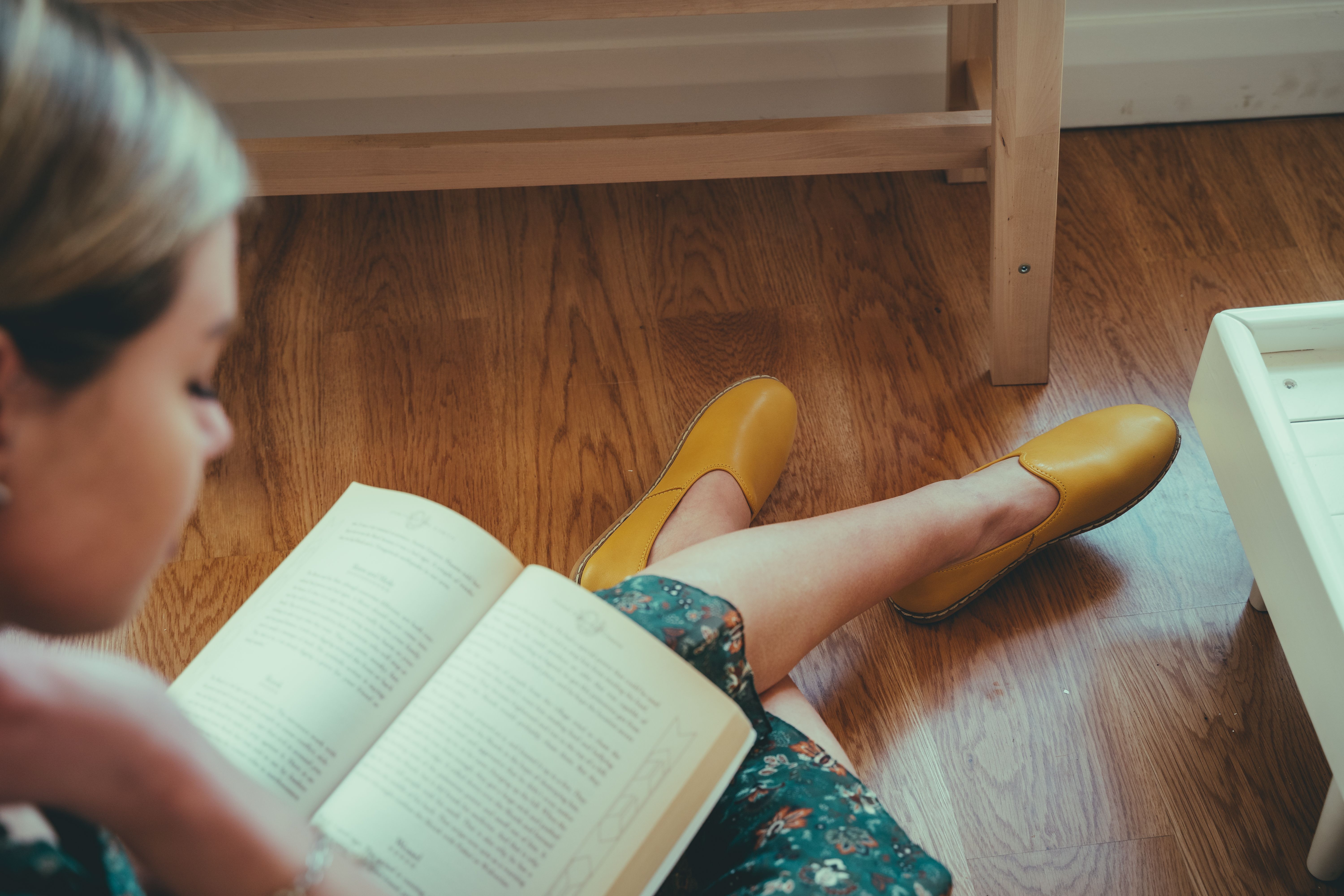 Woman sitting on a wooden floor reading a book, with yellow shoes and a floral dress, creating a cozy atmosphere.