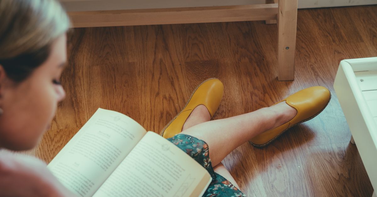 Woman sitting on a wooden floor reading a book, with yellow shoes and a floral dress, creating a cozy atmosphere.
