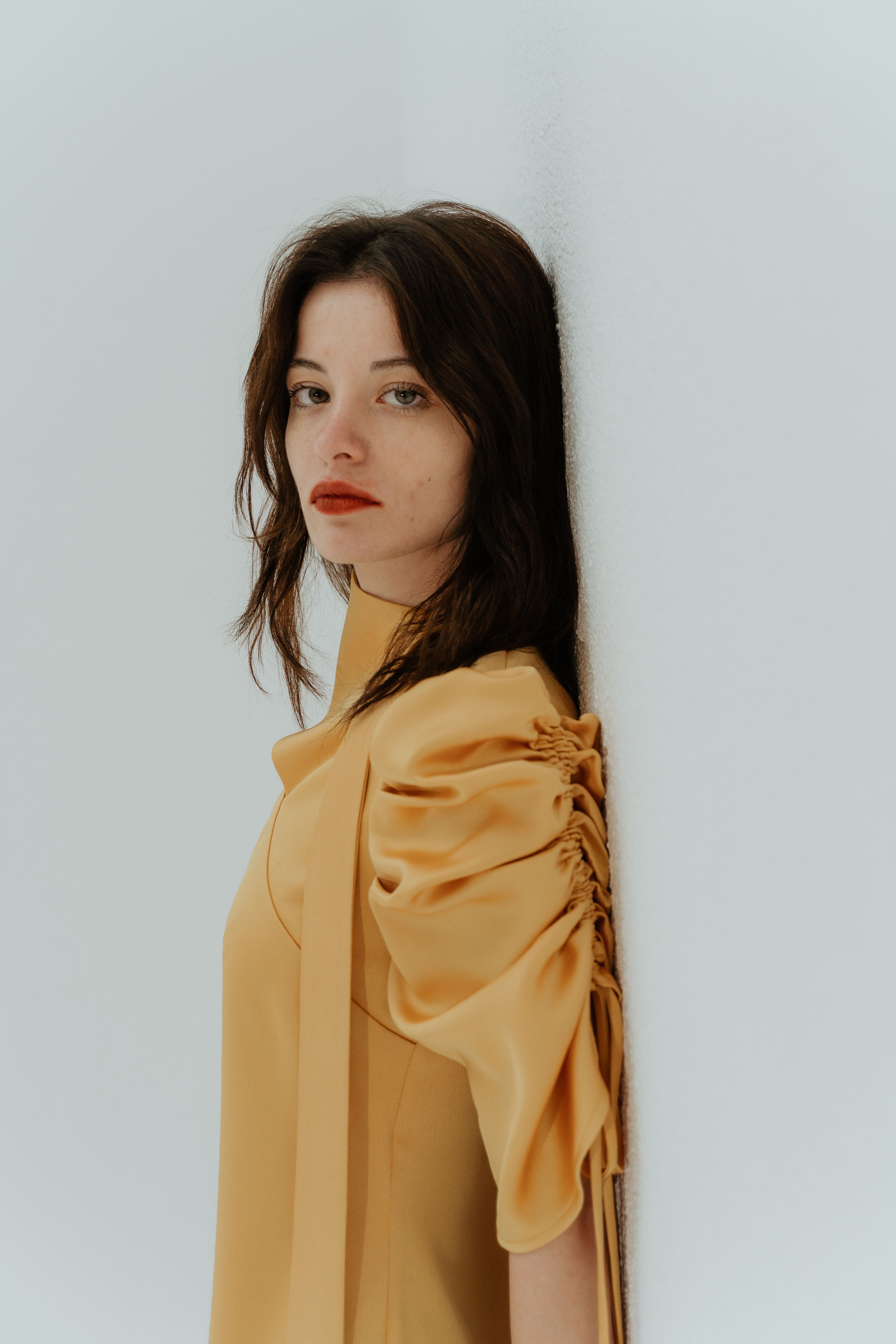 Stylish adult woman wearing yellow dress leaning against a white wall. Perfect for fashion and lifestyle themes.