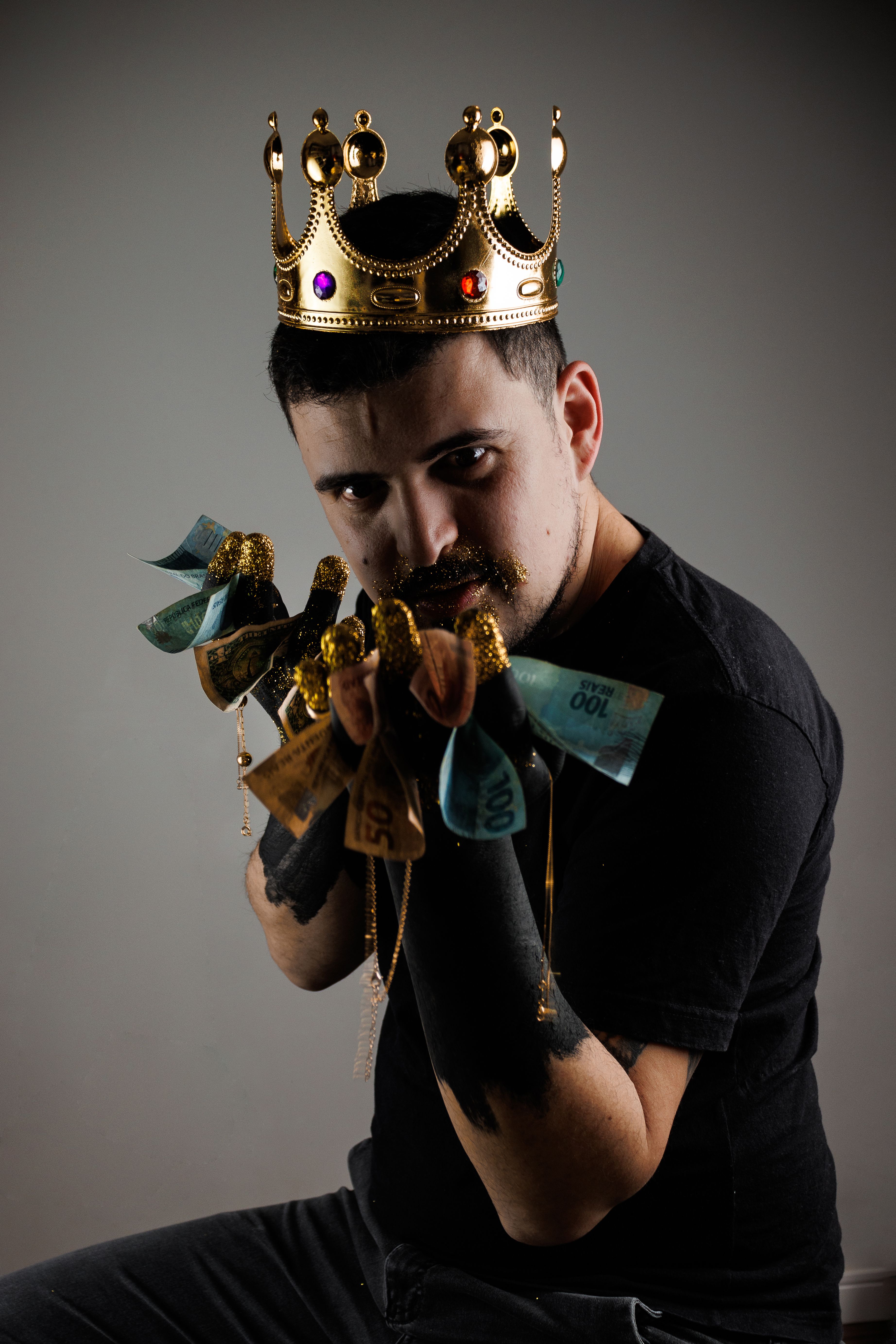 A man wearing a golden crown holds assorted glittering currency on a dark background.