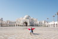A child runs joyfully in the grand courtyard of a Middle Eastern palace under a clear blue sky.
