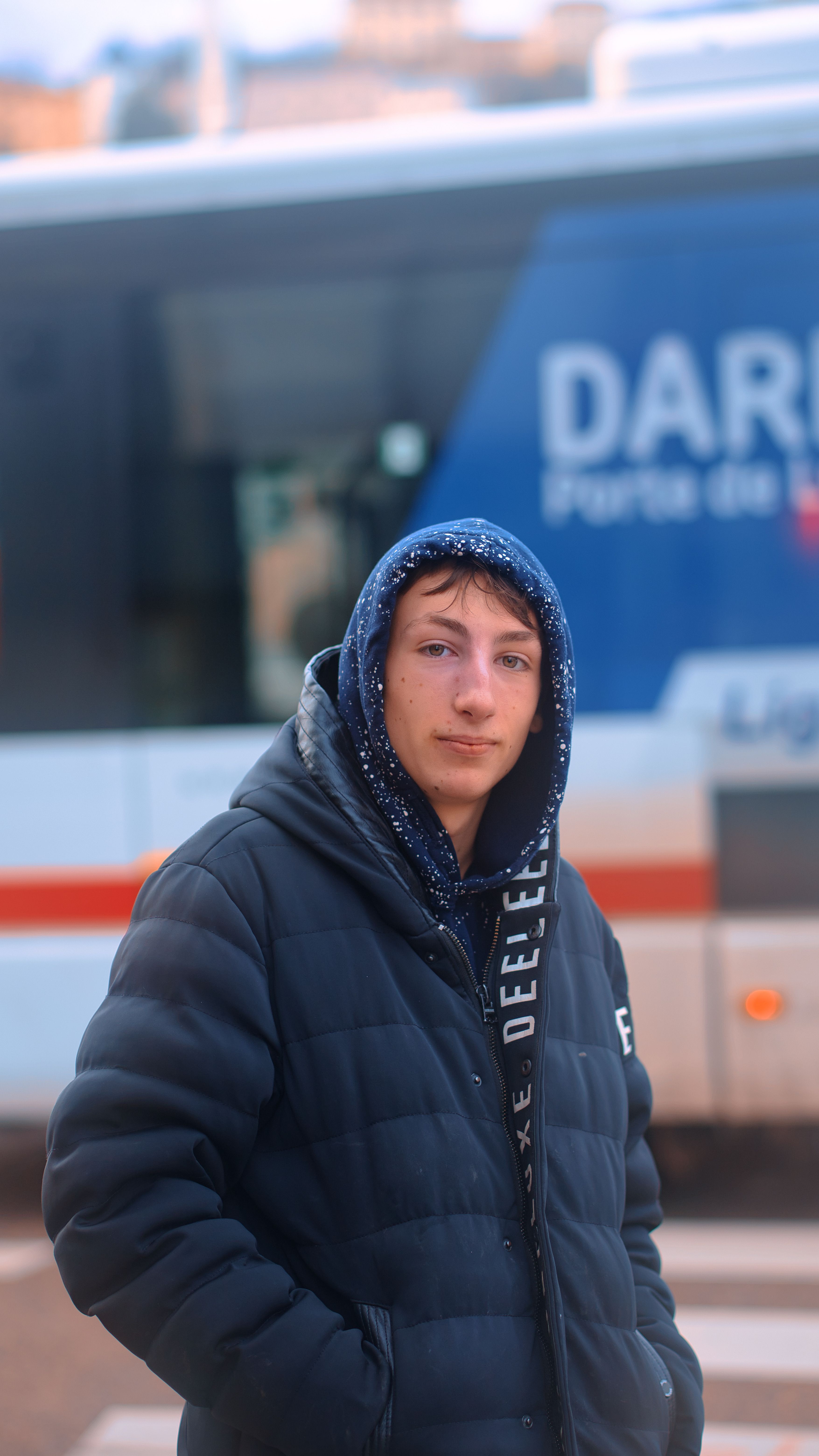 Young adult in a blue hoodie and puffer jacket standing in front of a bus.