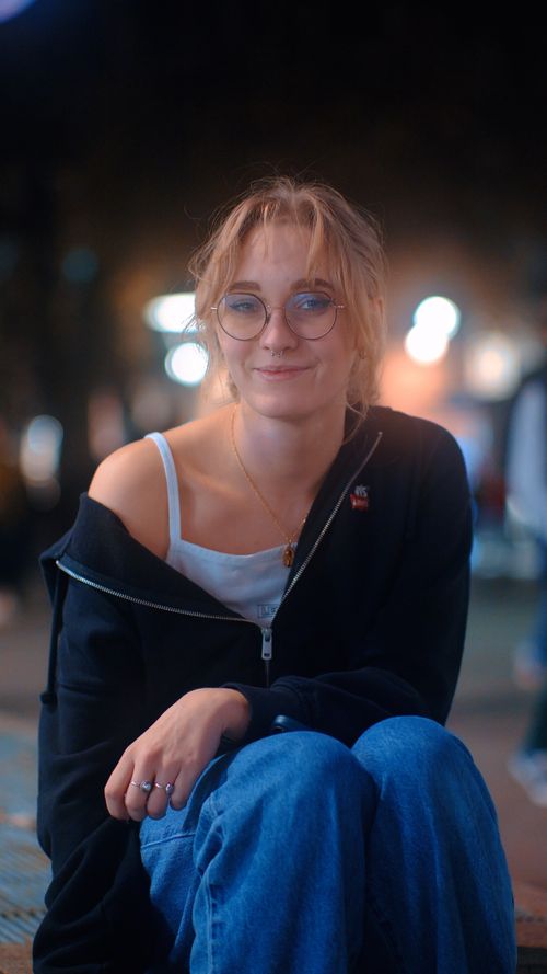 Free A young woman in casual fashion with glasses, smiling outdoors at night with urban lights in the background. Stock Photo
