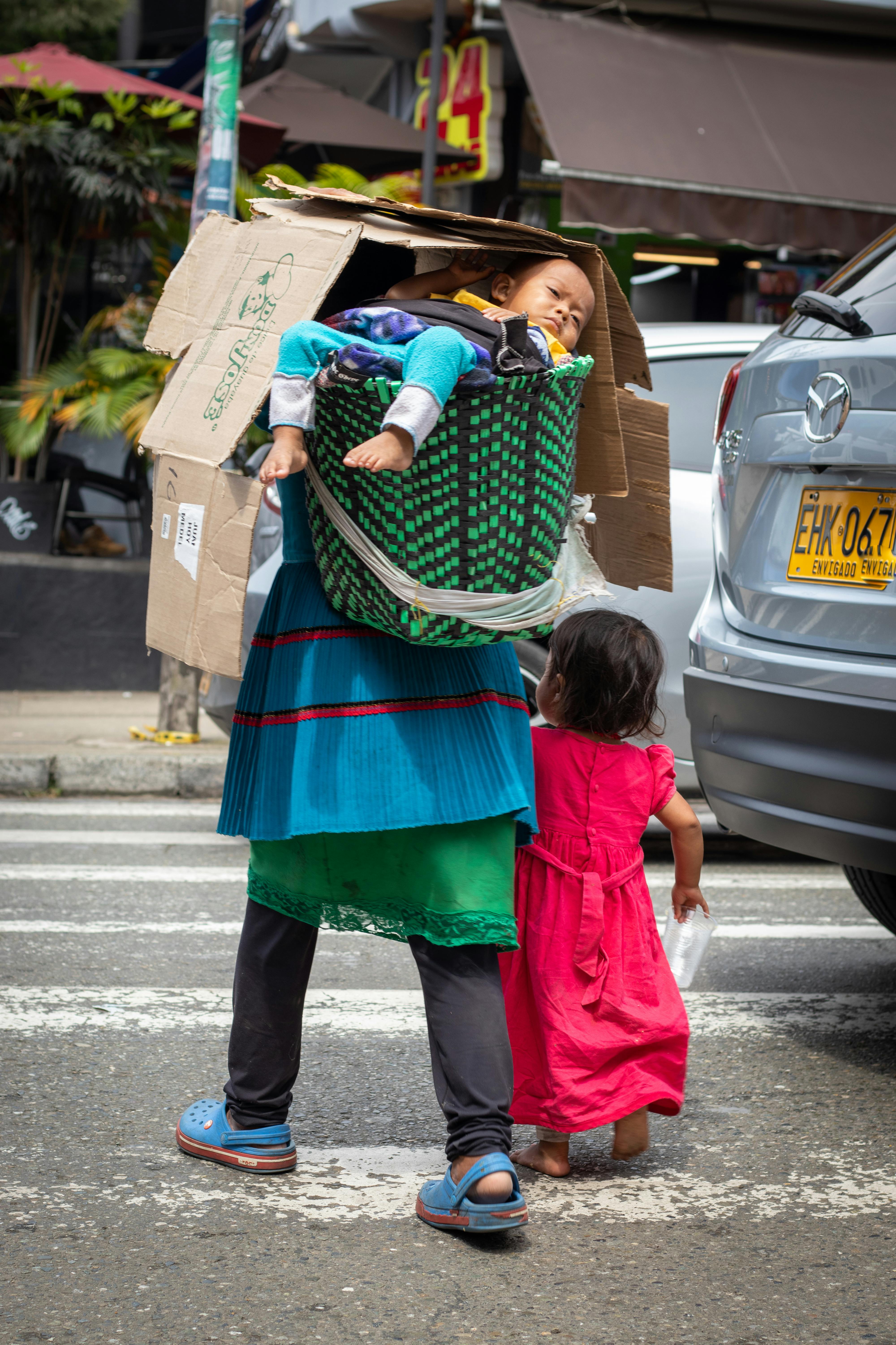 baby in a basket on the back of his mother leading her daughter by the hand