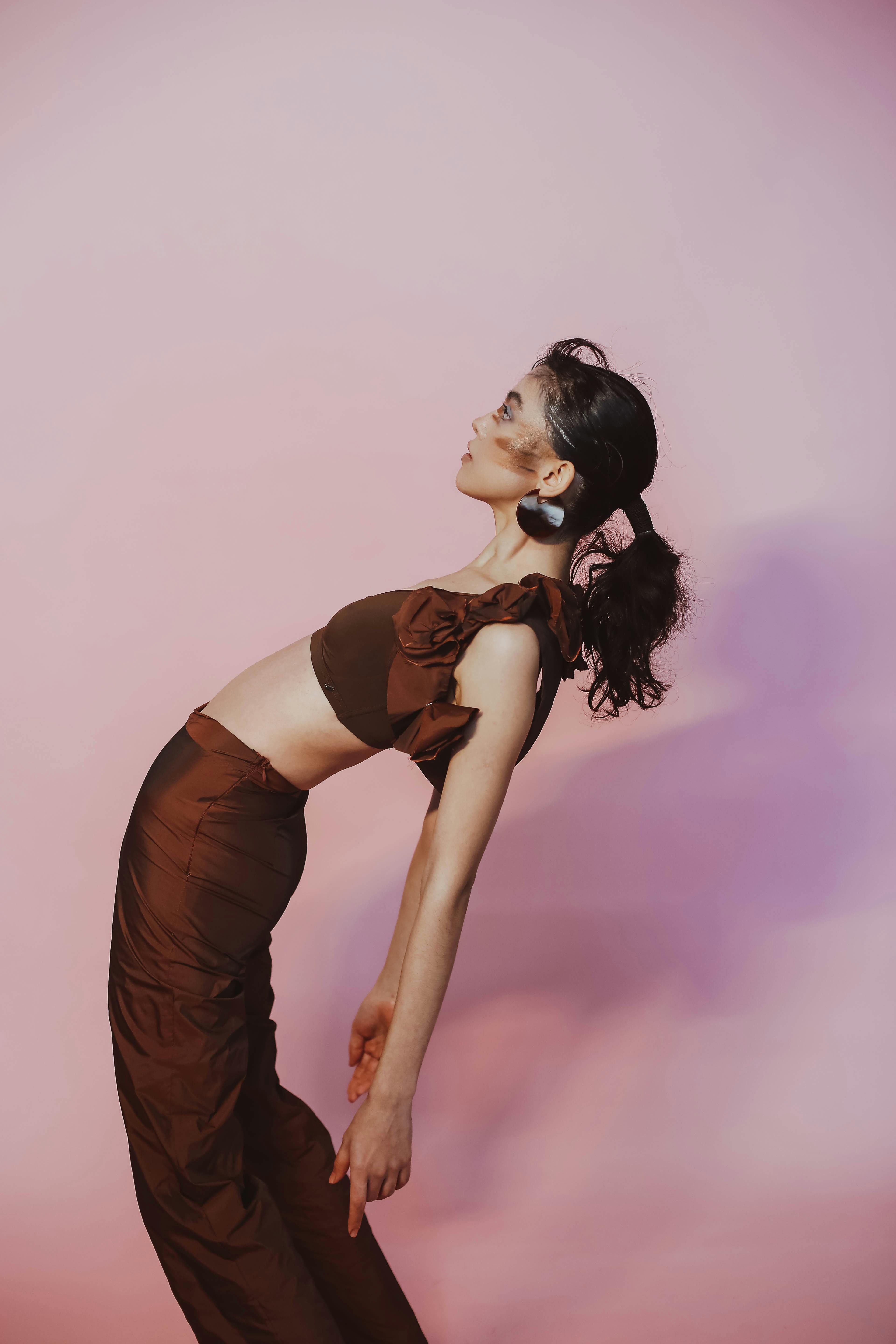 A stylish woman in brown fashion poses elegantly indoors against a pink background.