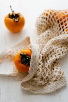 Close-up of vibrant persimmons in a textured mesh bag on a rustic white table.