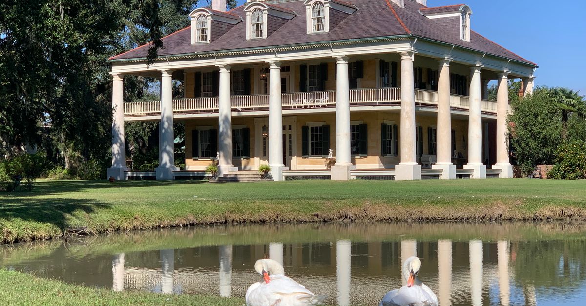 A stunning historic mansion reflected in a pond with serene swans in a summer setting.