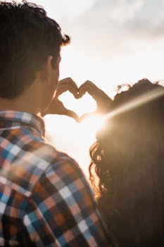 A couple in love creates a heart shape with their hands against a vibrant sunset backdrop.