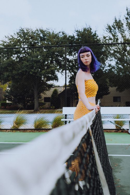 Free Woman with purple hair standing pensively on a tennis court in a yellow dress. Stock Photo