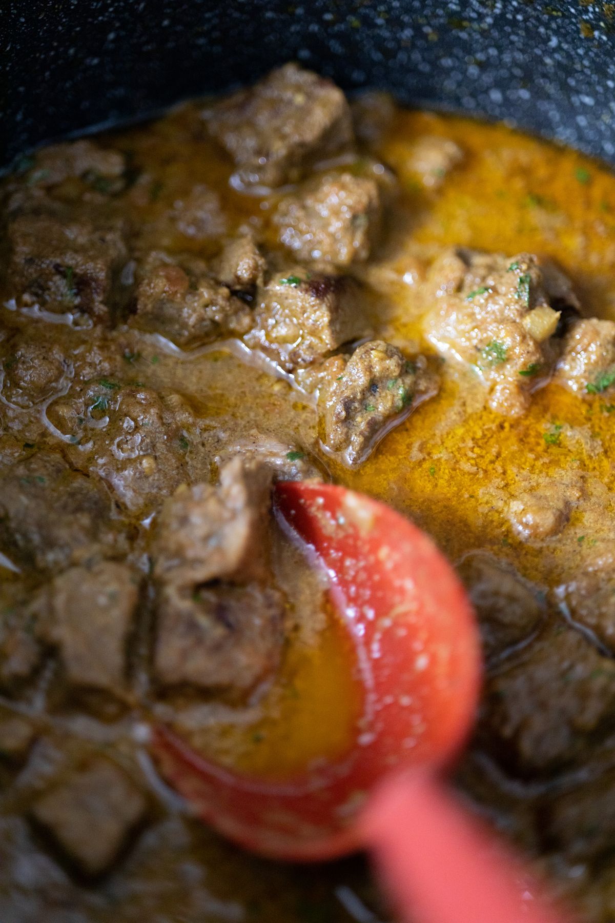 Close-up view inside a pot of freshly cooked beef stew with tender beef chunks in glossy brown gravy and a red silicone spoon