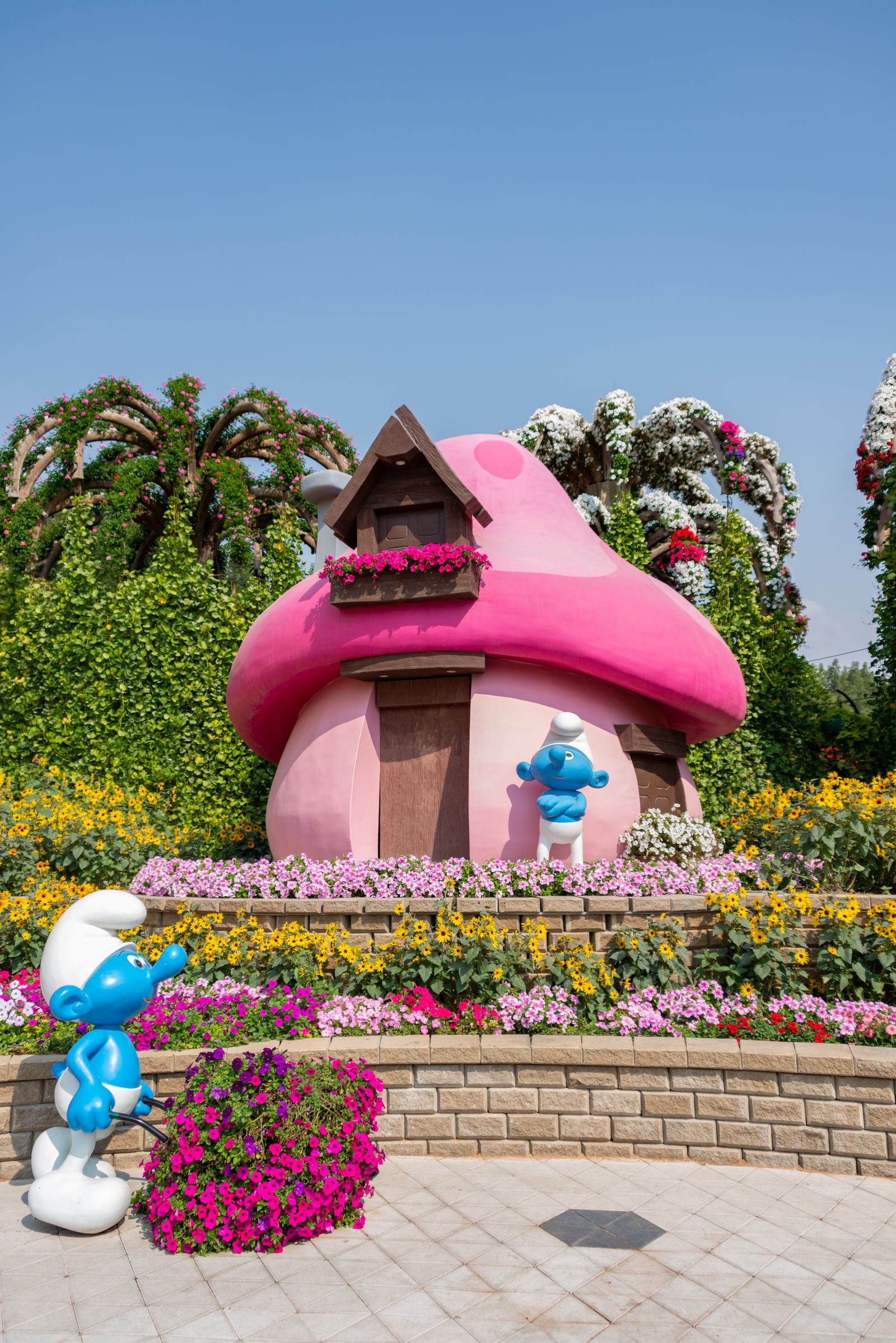 A pink mushroom-shaped Smurf house with blue figures surrounded by petunias and sunflowers at Dubai Miracle Garden.