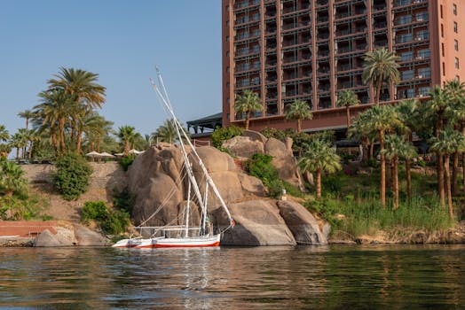 A serene sailboat rests on a calm river near boulders and lush palm trees, with a hotel in the background.