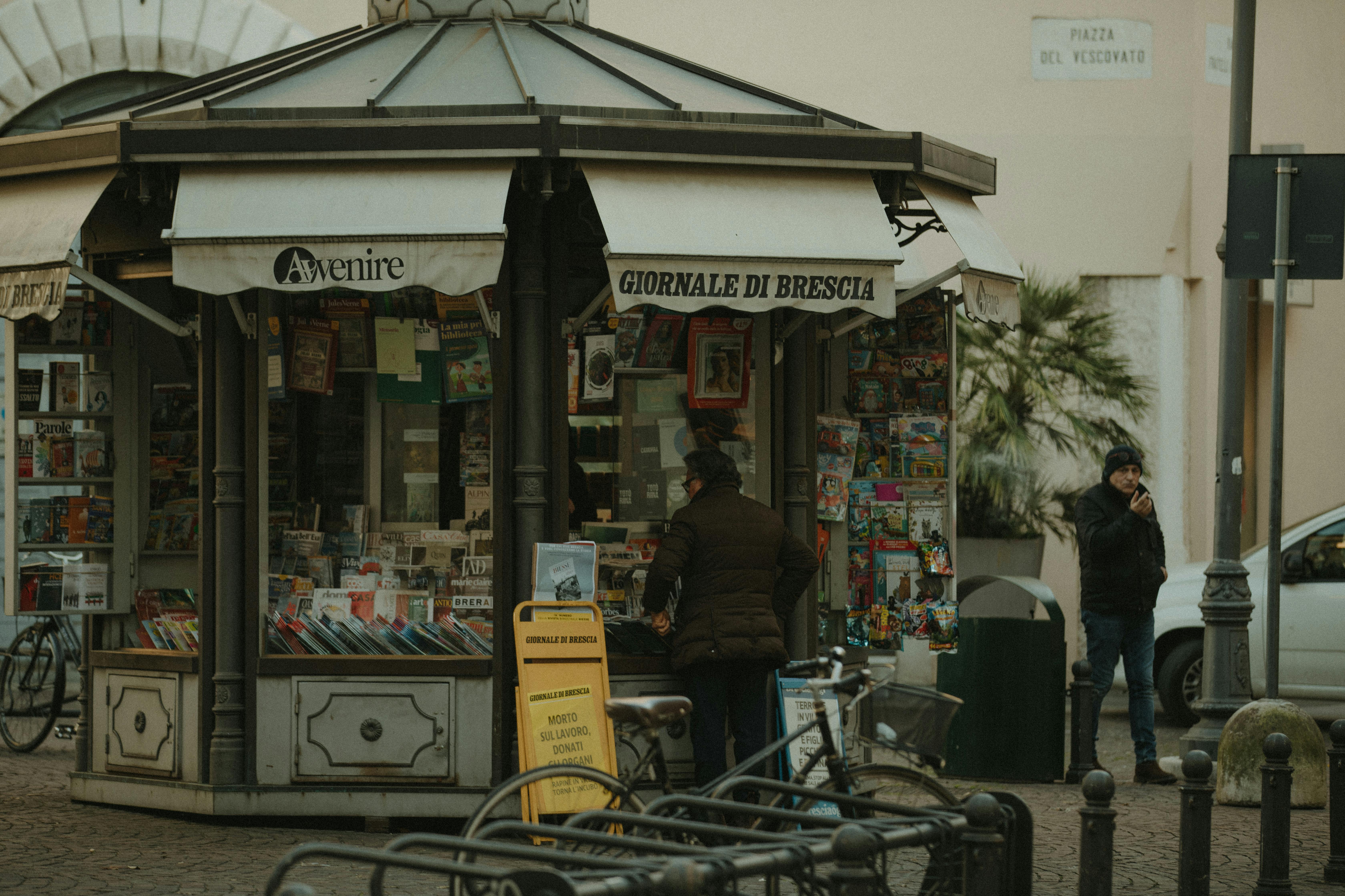 Free Street view of a newspaper kiosk in Brescia, Italy, featuring daily publications and passerby. Stock Photo
