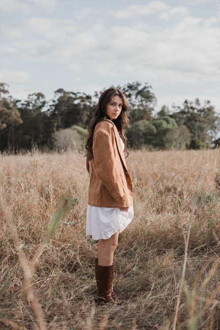 Young woman wearing a brown jacket and boots standing in a grassy field with trees.