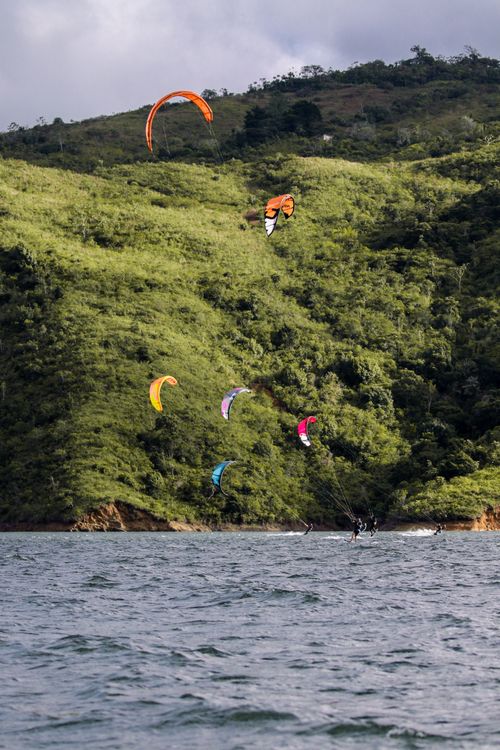 Free Vibrant kitesurfers glide across ocean waves with lush green hills in the backdrop. Stock Photo