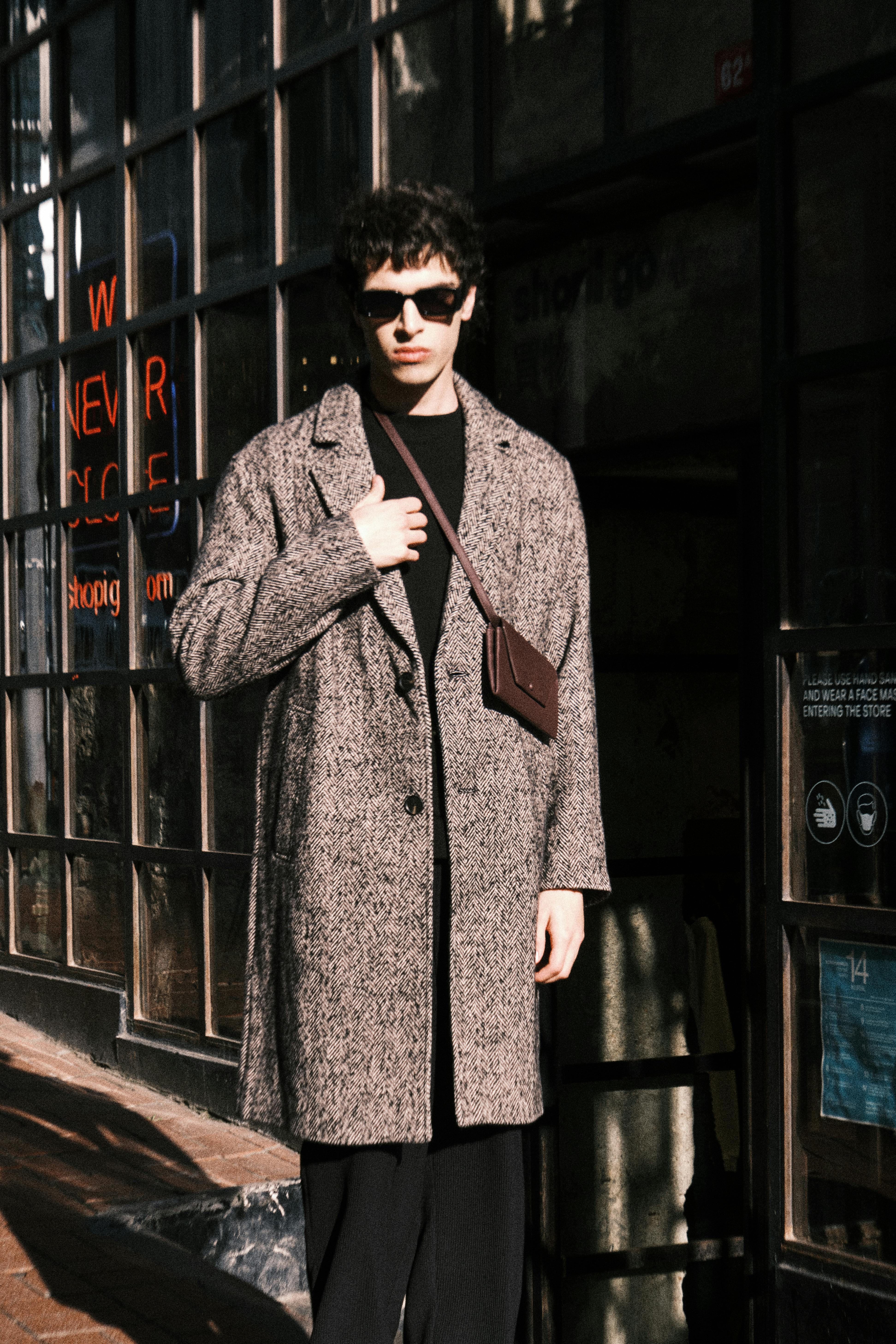 Stylish male model in coat, sunglasses, posing in urban area with window reflections.