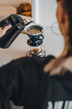Over-the-shoulder view of a woman barista pouring coffee with a gooseneck kettle.