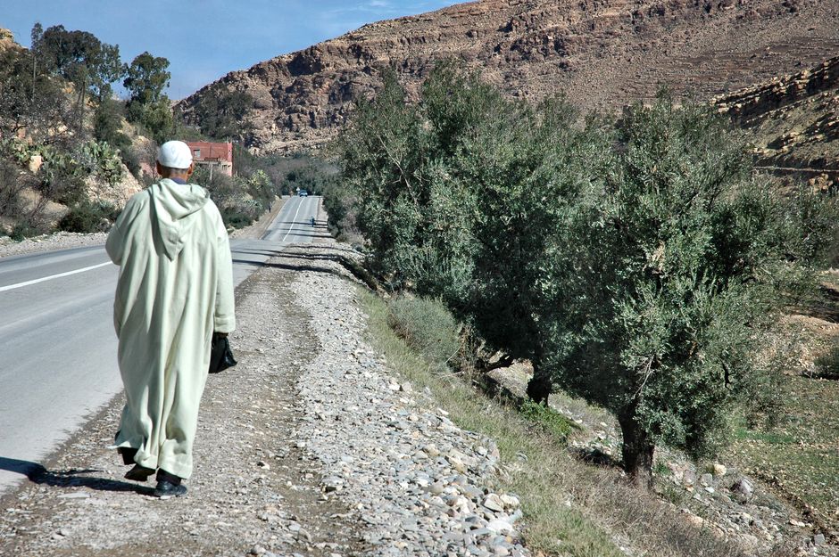 A man in traditional attire walks along a scenic road in Agadir, Morocco, surrounded by rocky landscapes.